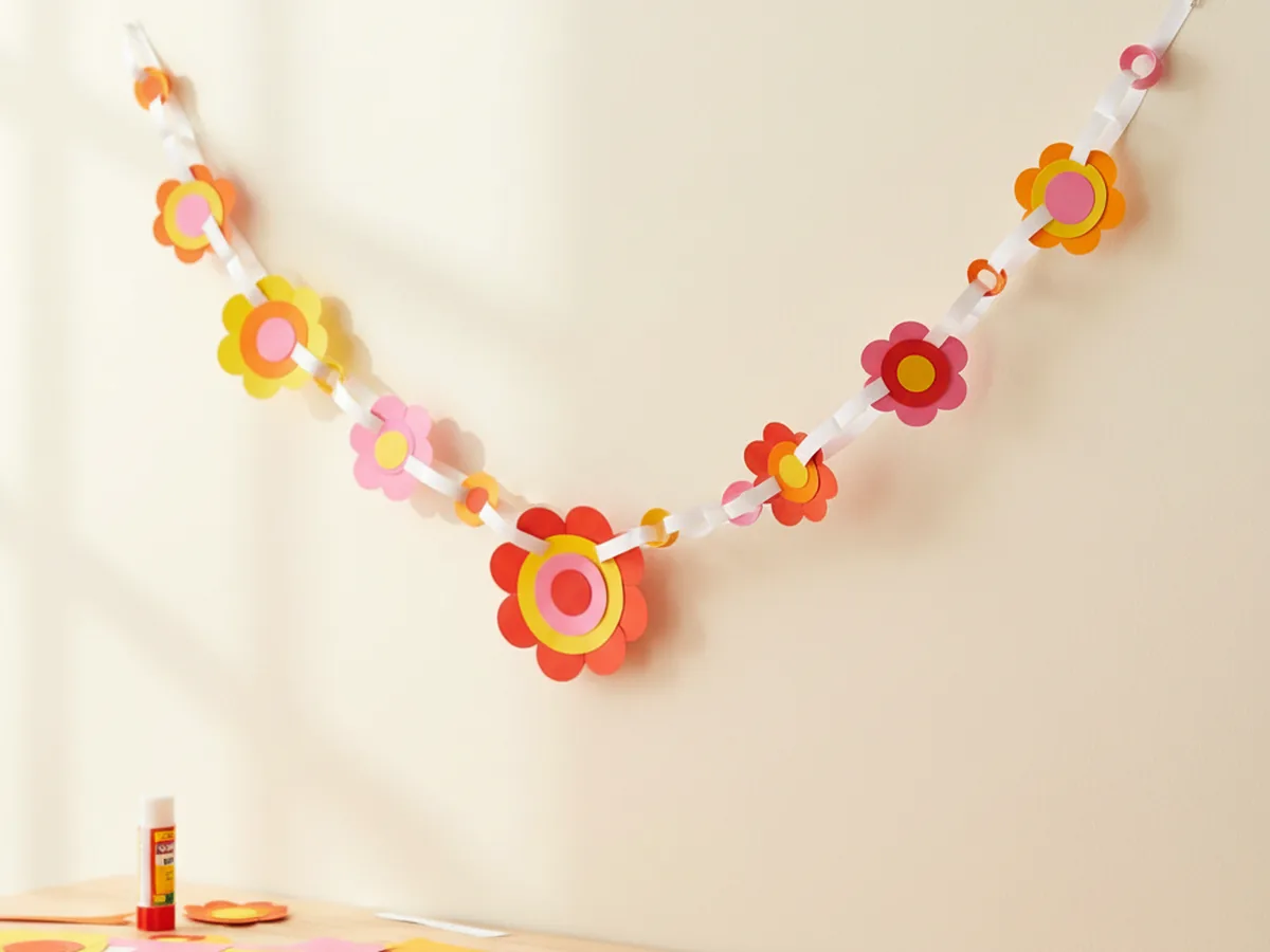 A handmade paper chain flower garland with small colorful paper flowers connected along a ribbon draped across a light wall