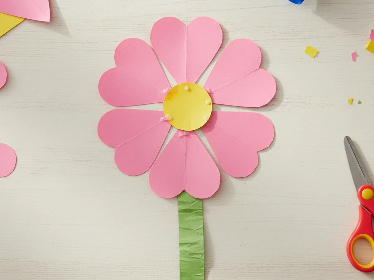 A handmade paper flower with pink heart-shaped petals arranged in a circle with a yellow center circle on a green stem