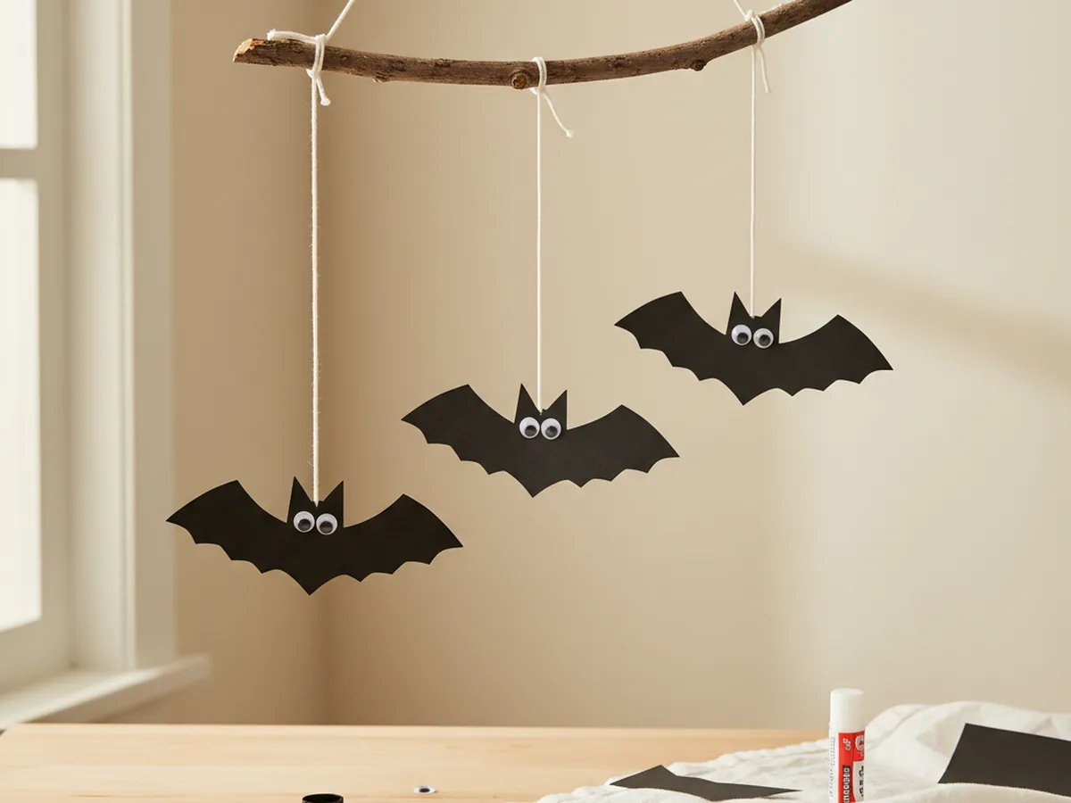 Handmade paper bat mobile with three black construction paper bat shapes hanging from strings on a wooden twig with googly eyes