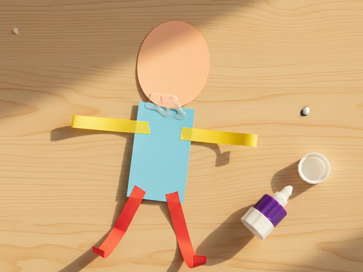 Assembled paper person body with oval head glued on top and four paper strip arms and legs attached behind, laid flat on a craft table