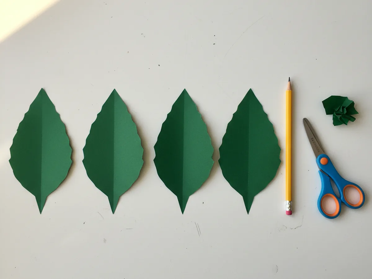 Five long pointed green construction paper leaves cut and laid out in a row on a white craft table next to a pencil and kid scissors, ready to be arranged into a poinsettia base