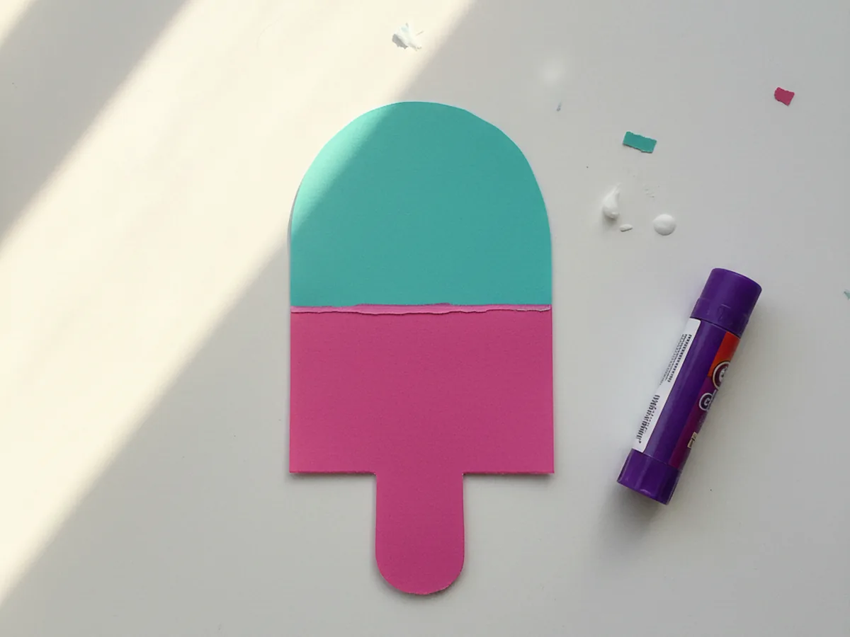 The pink paper popsicle body with a teal rounded-top paper layer glued over its upper half to create a two-tone two-flavor popsicle, resting on a white craft table with a glue stick nearby