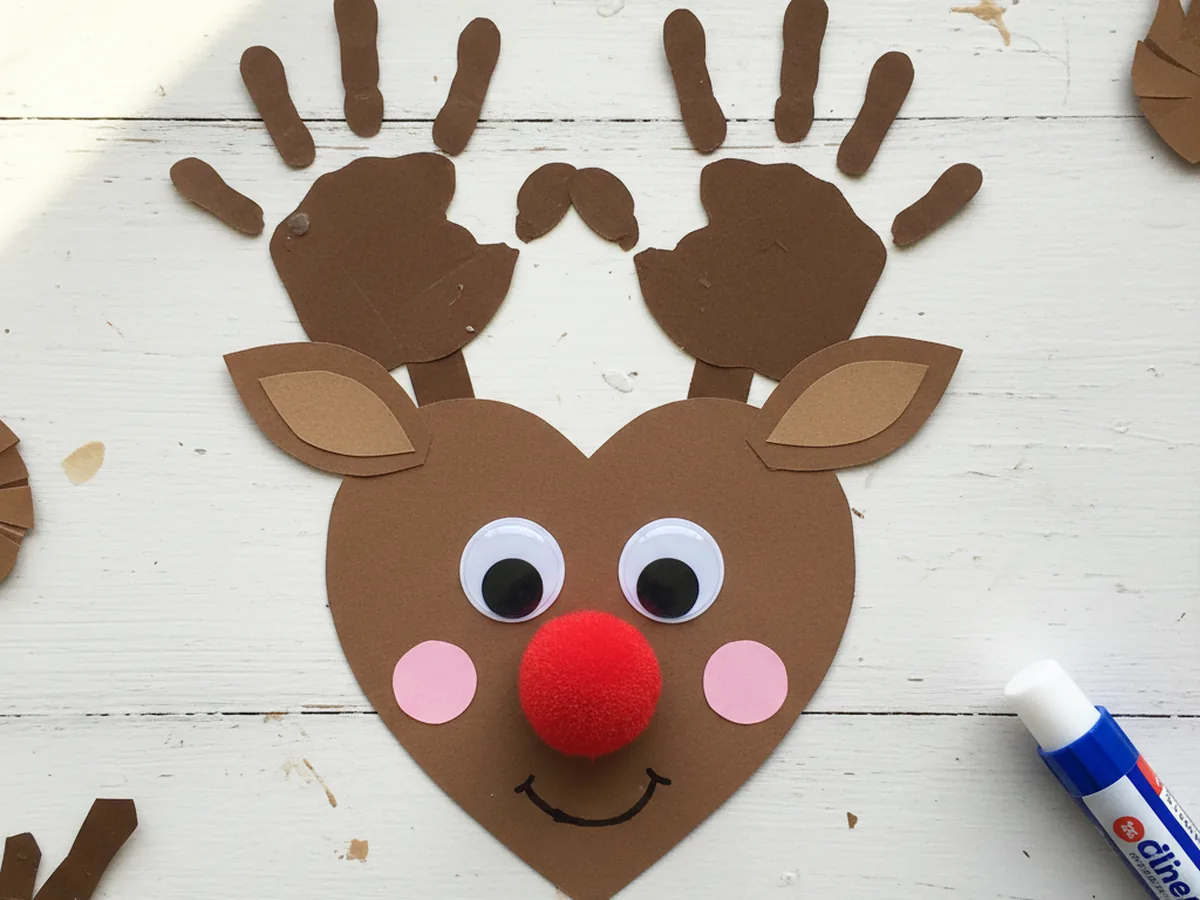 A finished paper reindeer craft with brown heart face, handprint antlers, googly eyes, fluffy red pom pom nose, tan inner ears, rosy cheeks, and a small marker smile displayed on a craft table