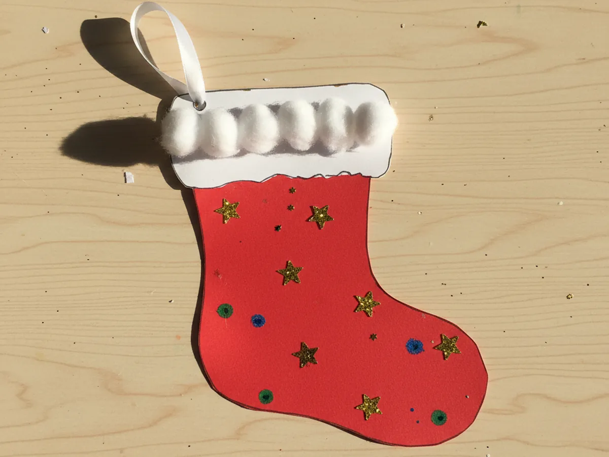 Finished handmade paper stocking craft made from red cardstock with a white cuff topped with fluffy cotton balls, decorated with gold star sequins and a thin white satin ribbon loop for hanging