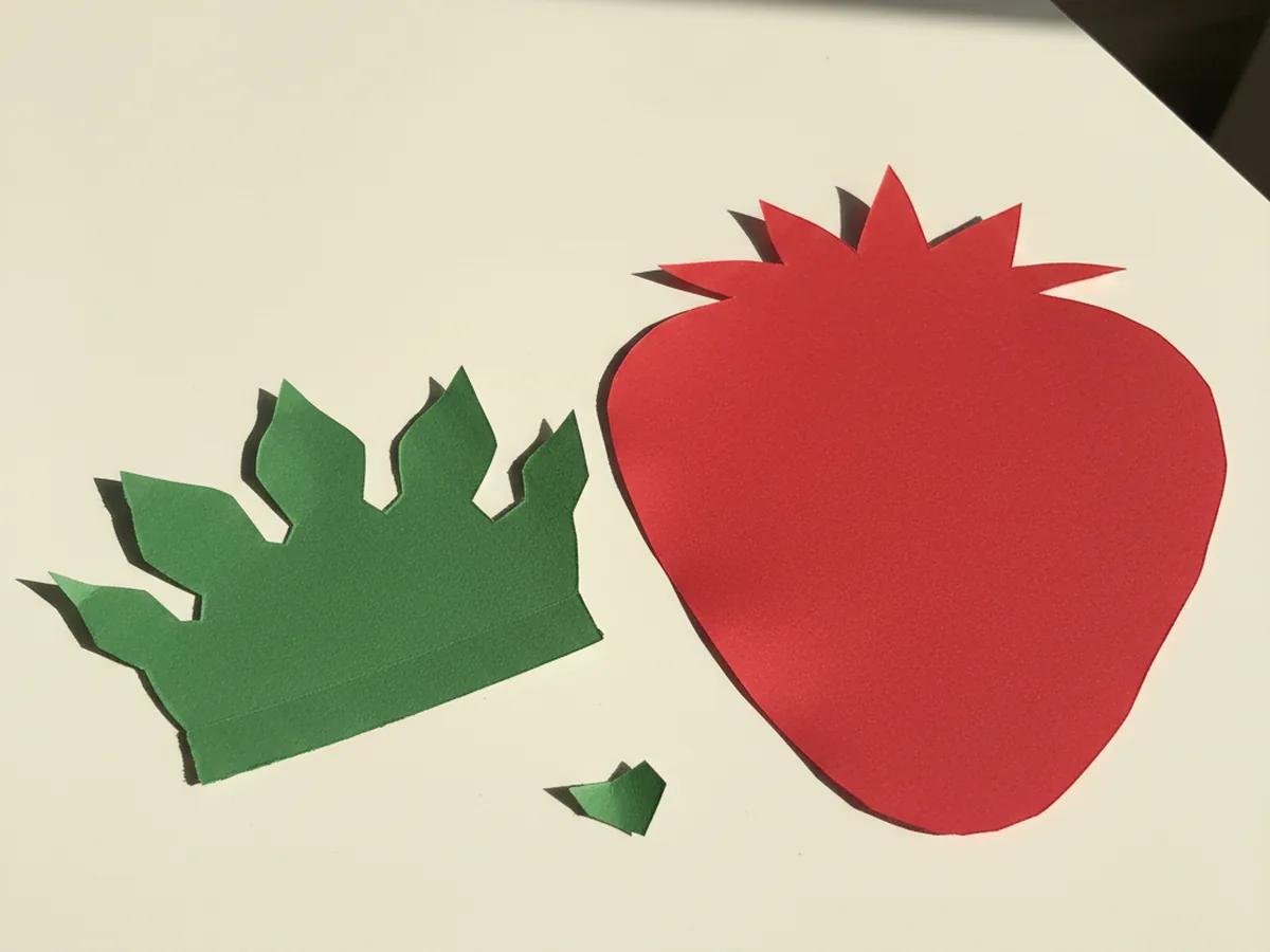 A green construction paper crown with five or six gently pointed leaves arranged in a star shape, freshly cut and laid next to the red strawberry body on a white craft table