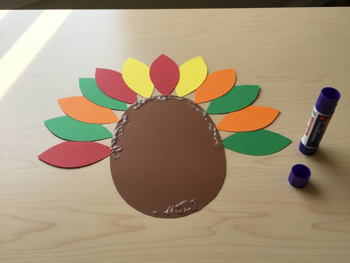 Colorful paper feathers fanned out and glued behind a brown construction paper turkey body on a white table