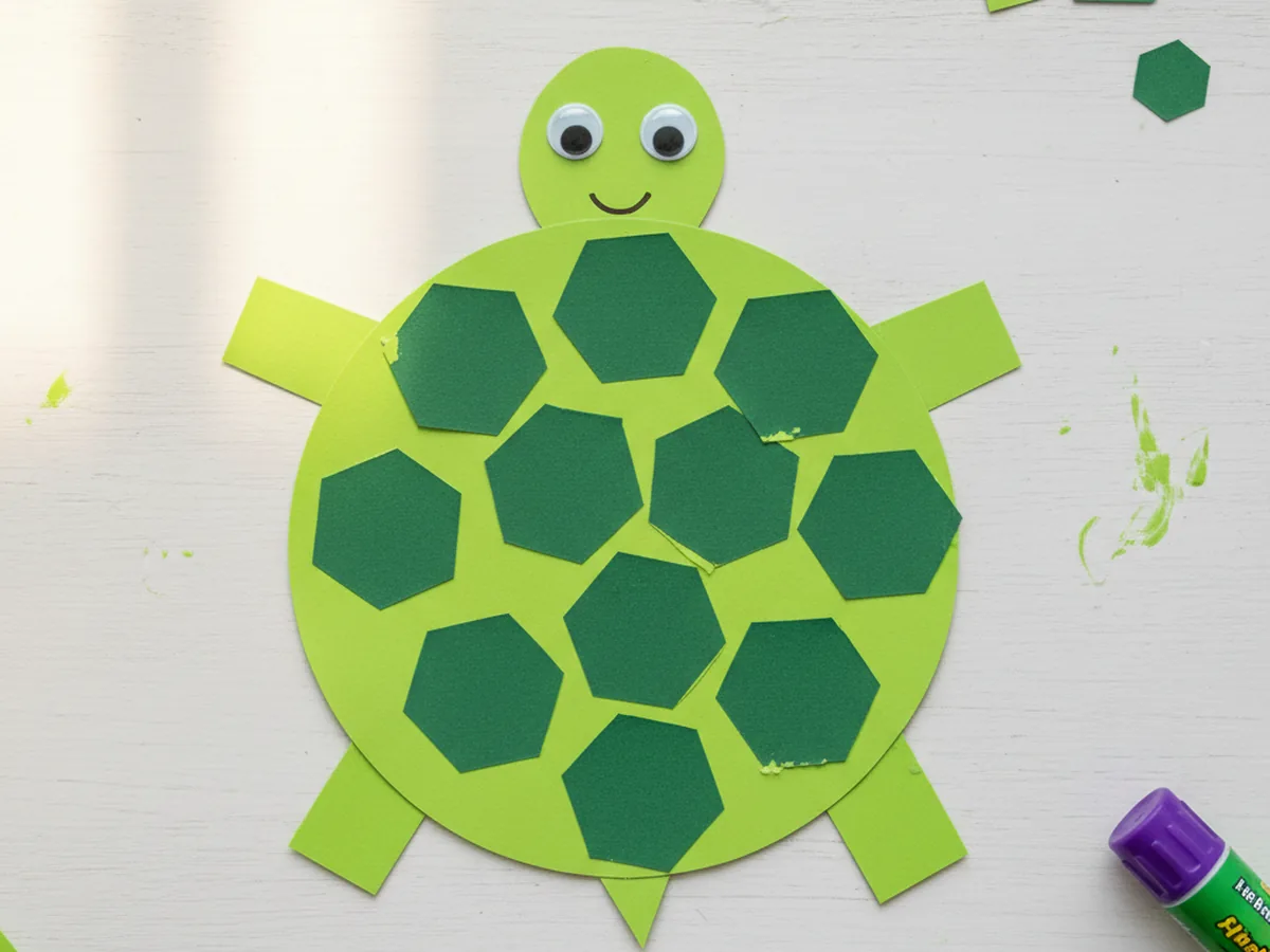 Finished green paper turtle craft with a hexagon-patterned shell, googly eyes, and a smiling face on a white craft table
