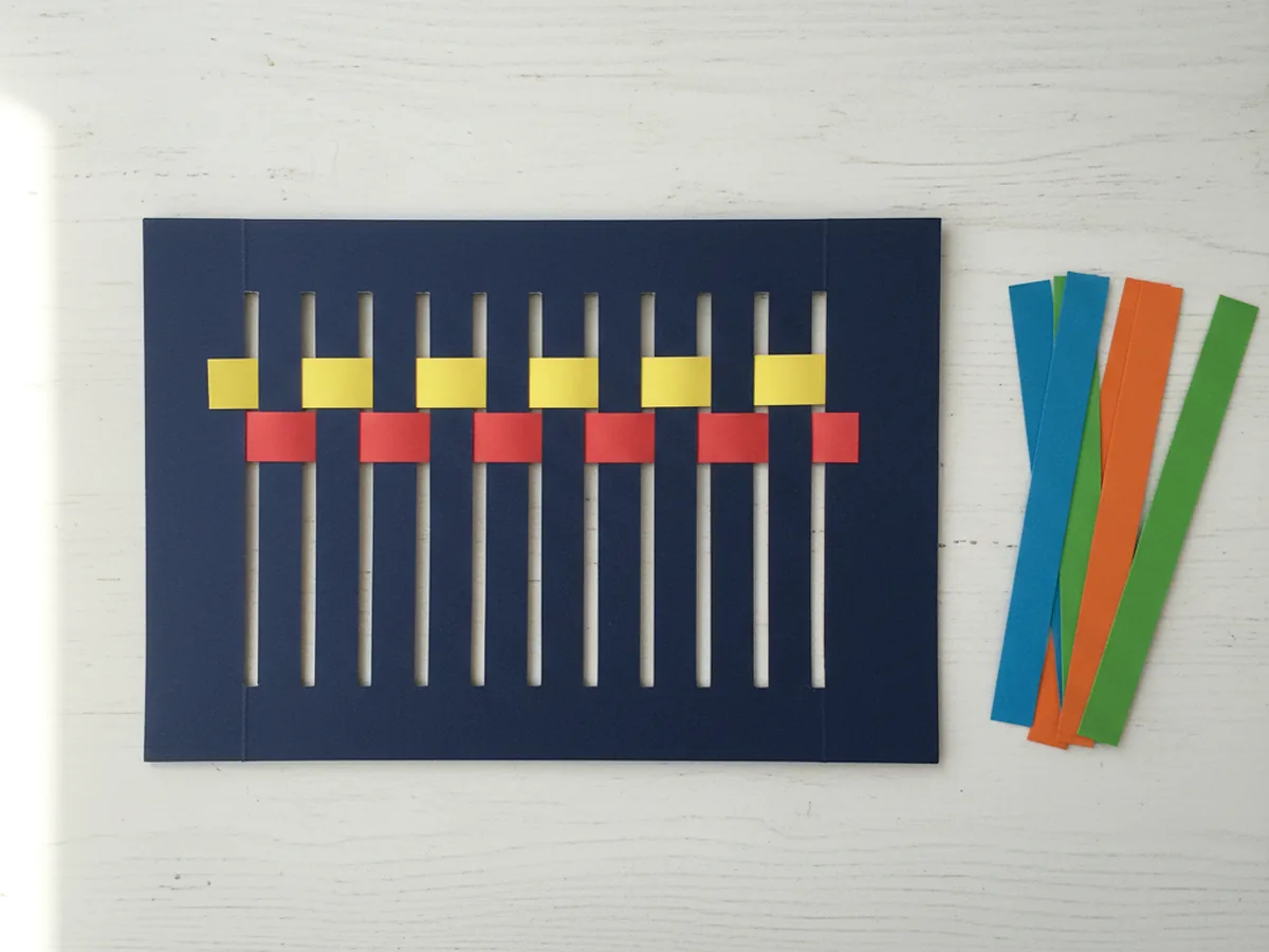 A red construction paper strip woven into a dark blue paper loom under and over each section, snug against a yellow strip already in place for the paper weave craft