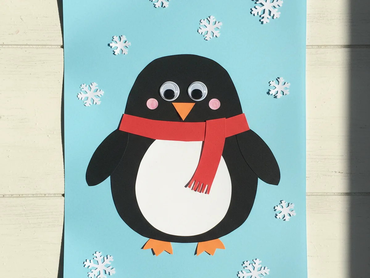The finished penguin paper craft with a small red scarf around its neck and white paper snowflakes scattered around the light blue background