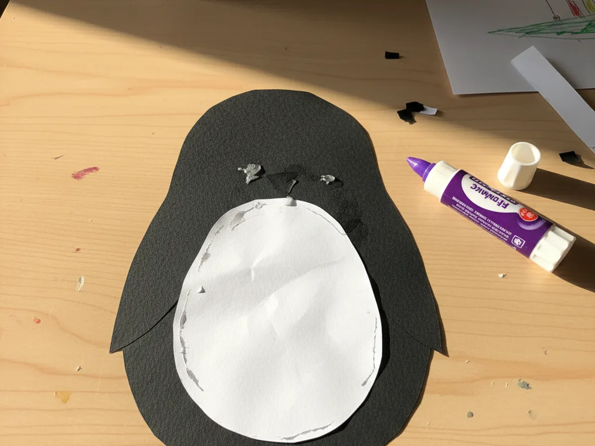 A smaller white paper oval glued onto the front of the black penguin body, sitting on a craft table to show the contrast between black and white
