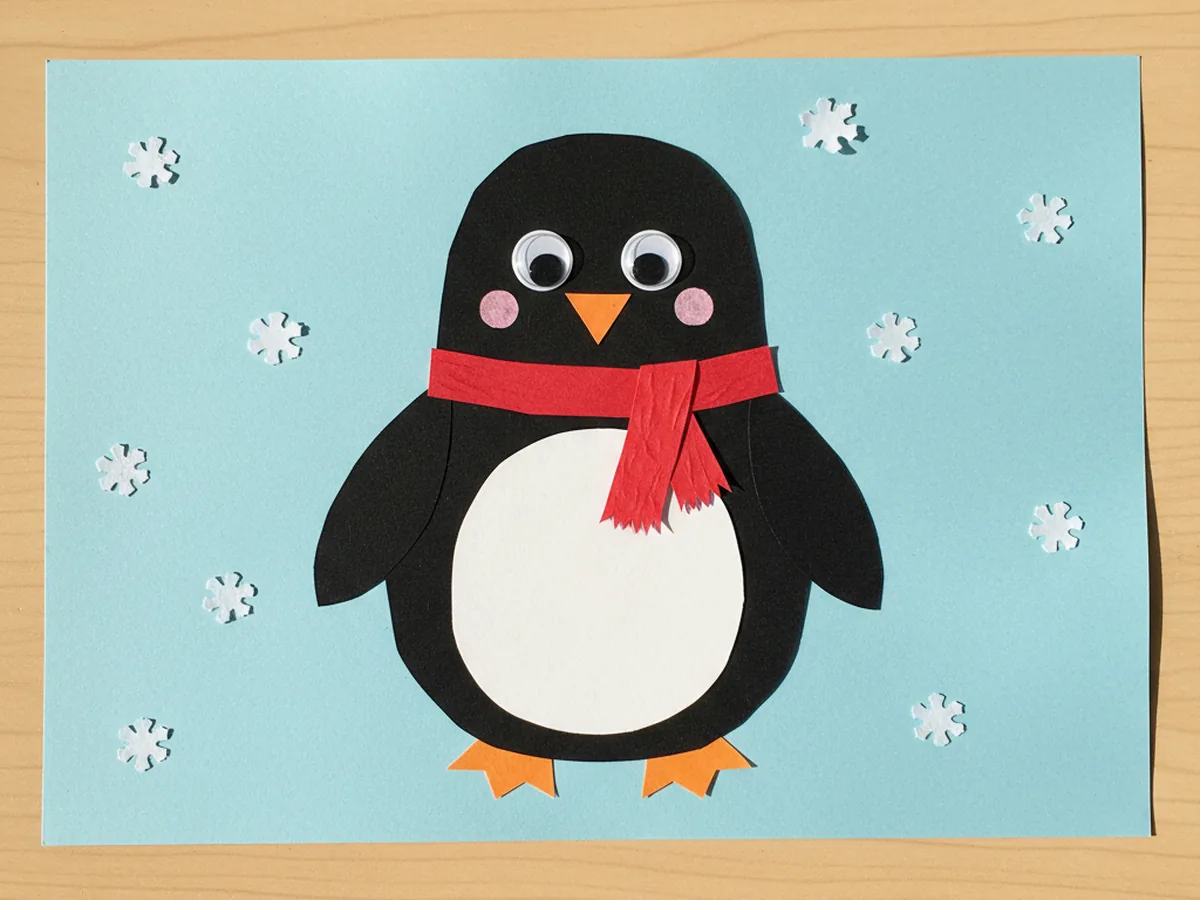 Finished penguin paper craft with black body, white belly, orange beak and feet, googly eyes, and a tiny red scarf on a light blue snowy background