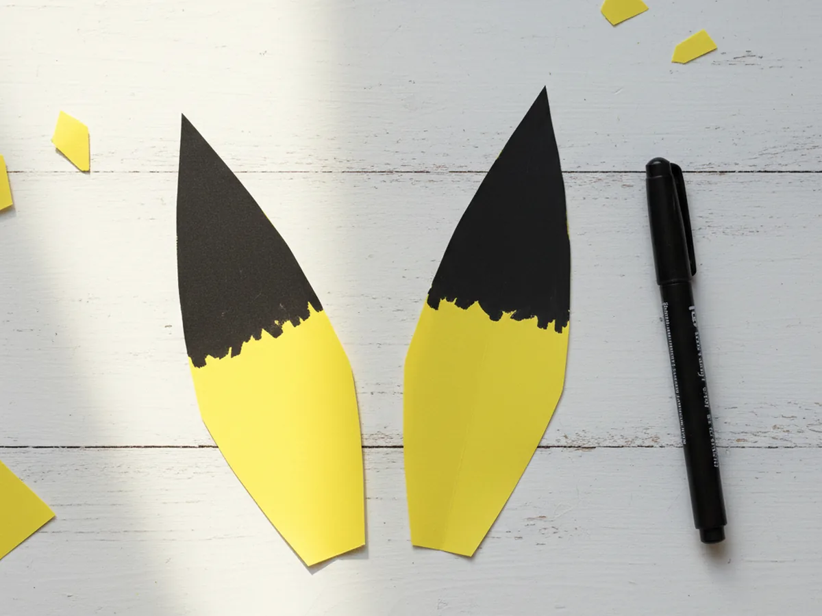 The two yellow Pikachu paper ears with the top third of each ear colored solid black to create the classic Pikachu ear tips, lying on a white craft table