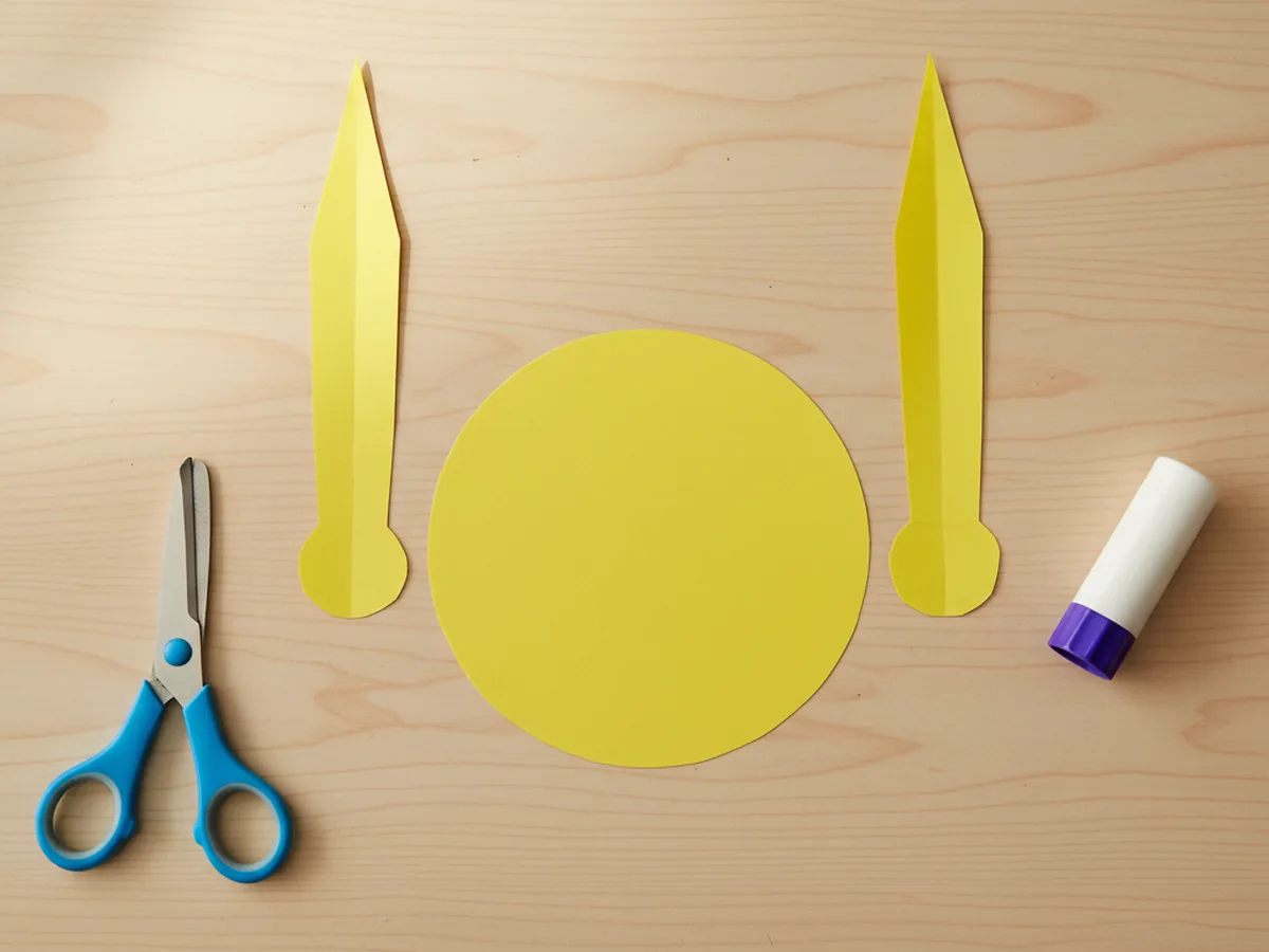 Two tall pointed yellow paper ear shapes for the Pikachu pokemon craft paper laid out next to the large round yellow head on a white craft table