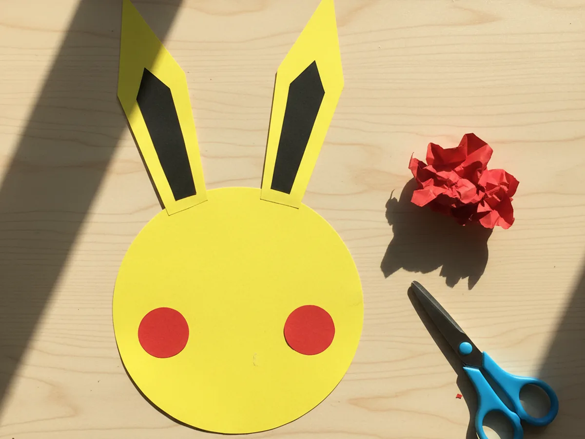 The yellow Pikachu paper craft with two small red paper circle cheeks glued onto the lower half of the face, one on each side, on a white craft table