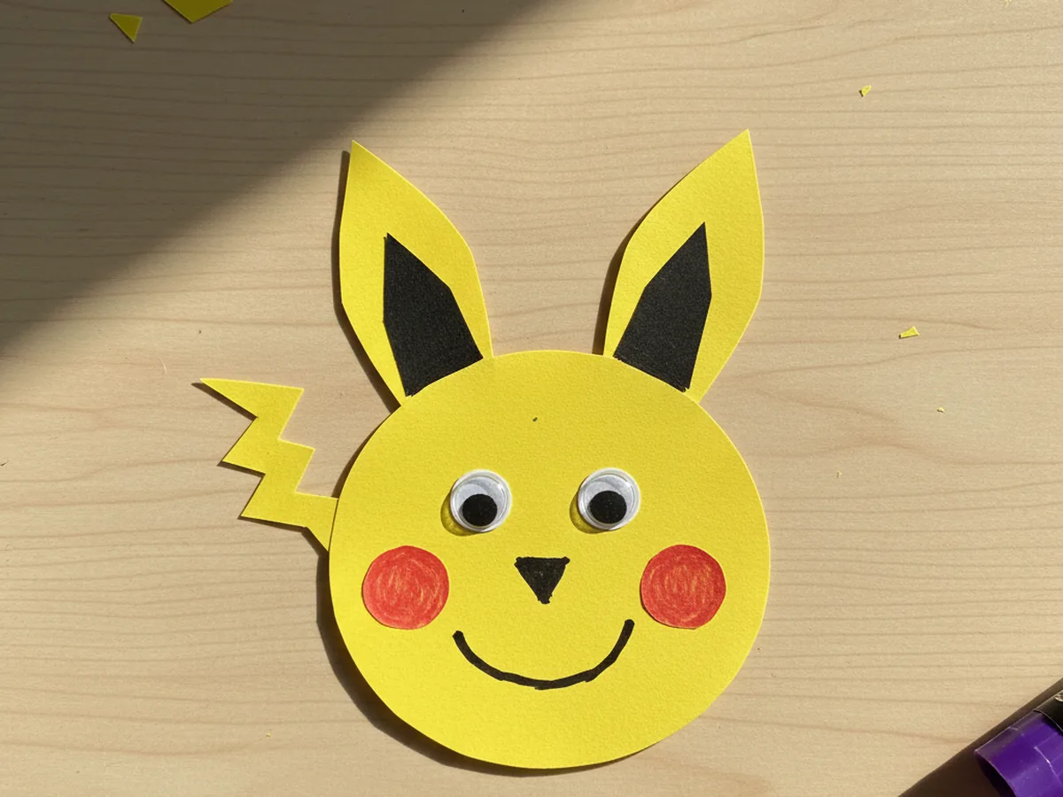 Finished yellow Pikachu pokemon craft paper with long pointed ears, red cheek circles, googly eyes, and a friendly smile on a white craft table