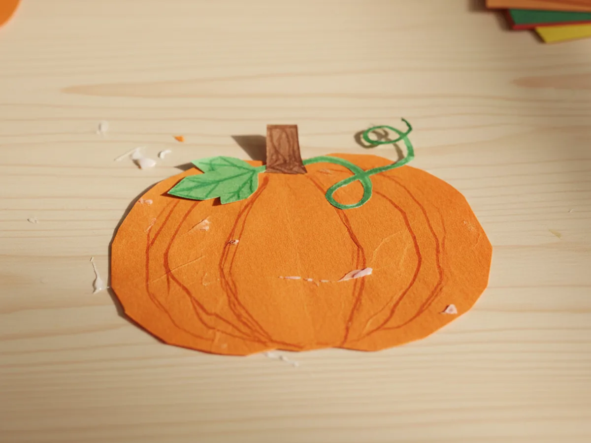 A handmade classic round orange construction paper pumpkin with a brown stem and a small green leaf and curly vine on a wooden craft table