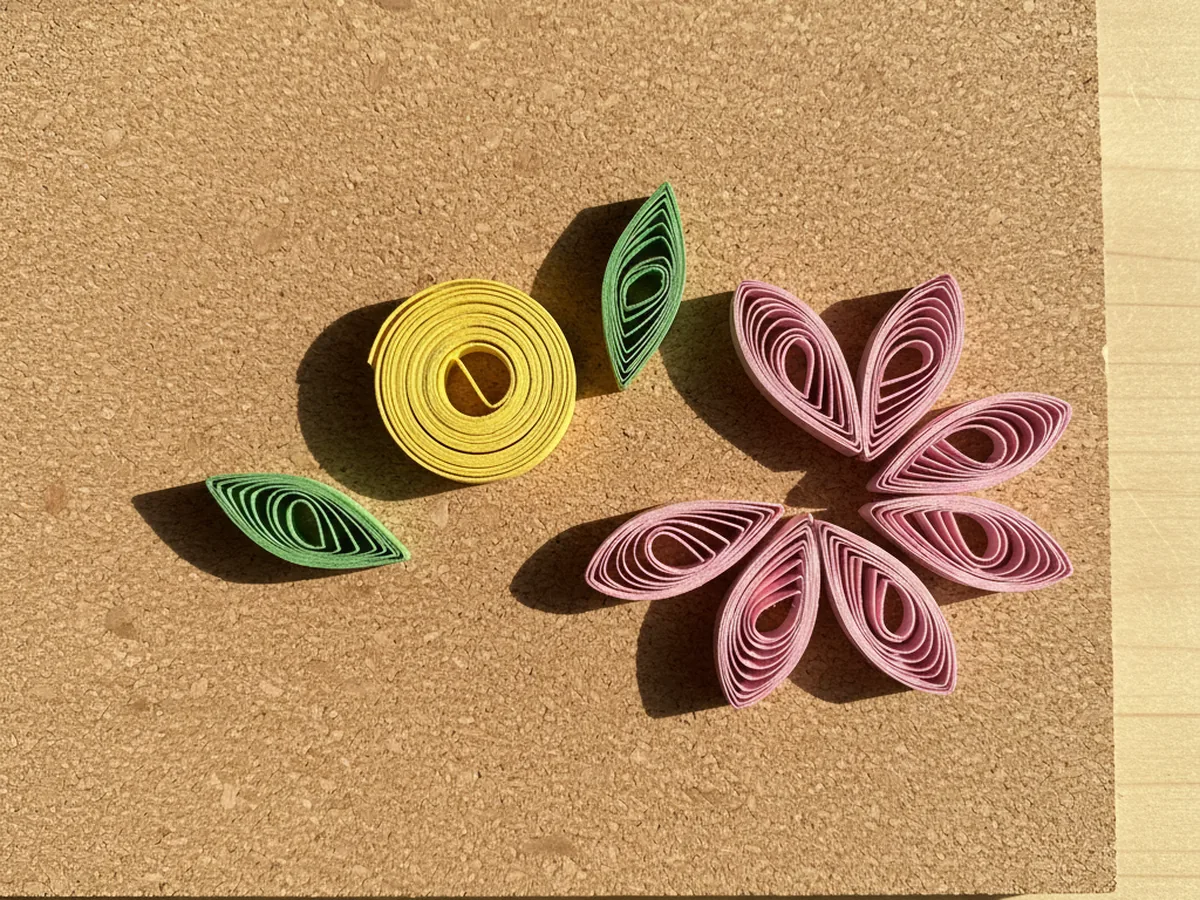 A small tight yellow quilling coil sitting next to two green marquise leaf shaped quilling coils with pointed ends, all arranged on a tan cork board with five pink teardrop petals nearby