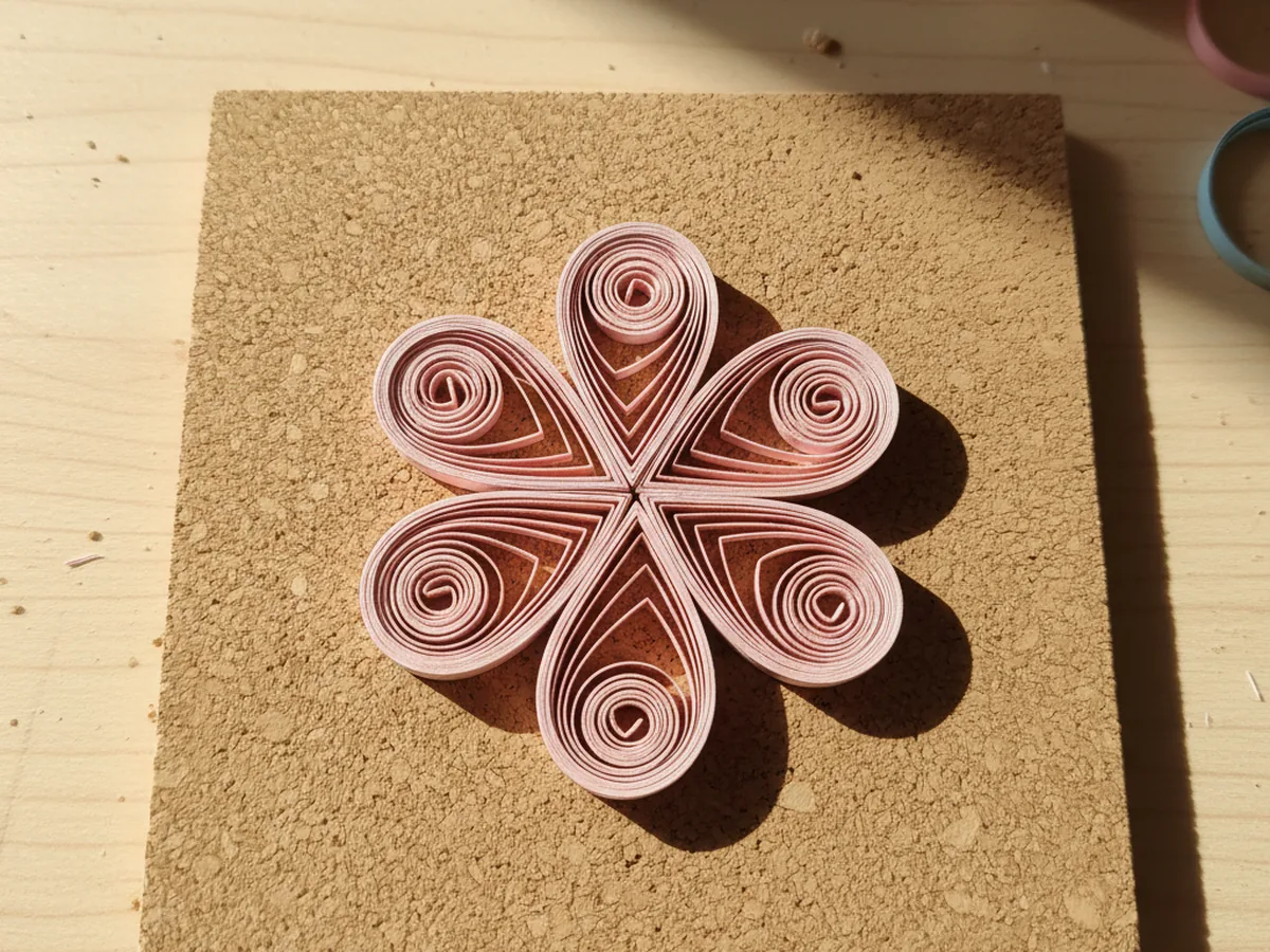 Five small pink quilling paper coils pinched into teardrop petal shapes arranged in a circle on a tan cork board with the pointed ends facing toward the empty center