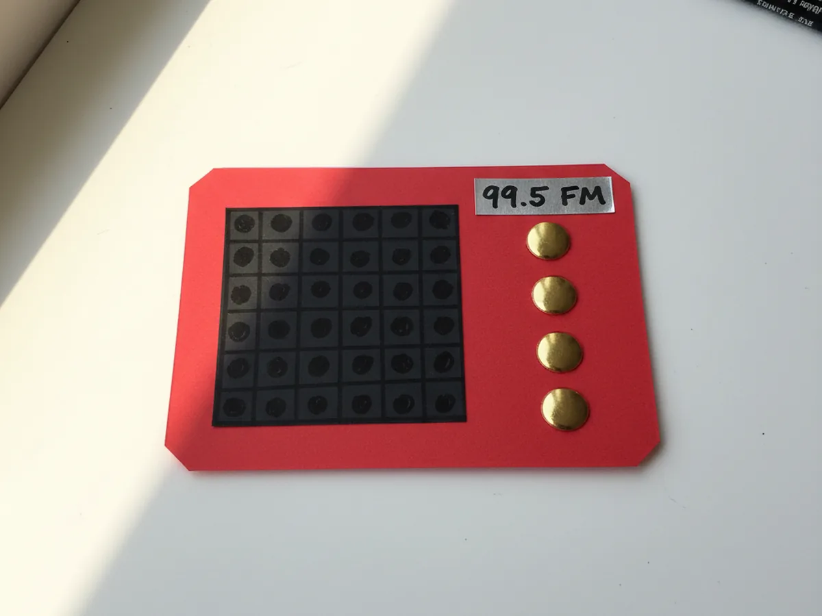 The red radio body with the black speaker grille and silver 99.5 FM frequency display now showing three shiny gold brass brad fasteners pushed vertically through the right side of the cardstock to serve as volume and tuning knobs