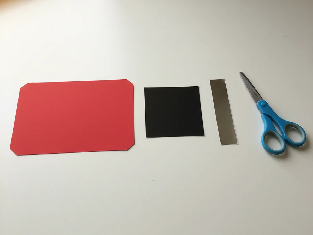 A small black cardstock square of about three inches and a thin silver metallic cardstock rectangle of about three inches wide by one inch tall, freshly cut and placed next to the red radio body rectangle on a white craft table