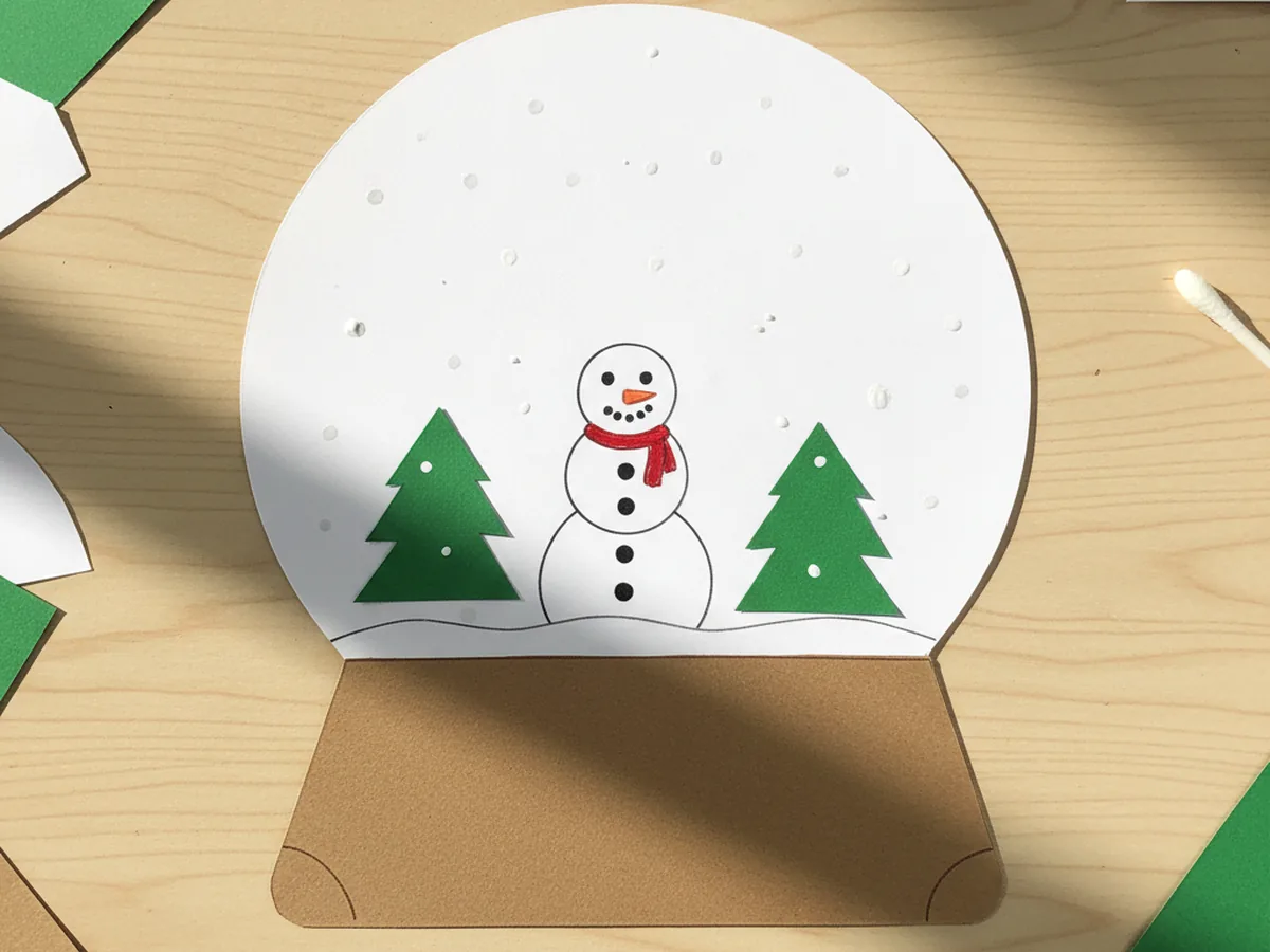 Finished snow globe paper craft with a white circle dome on a brown rectangle base, holding a tiny paper snowman and two small green construction paper trees, with white painted snow dots dotted across the inside of the globe, sitting on a light wood craft table