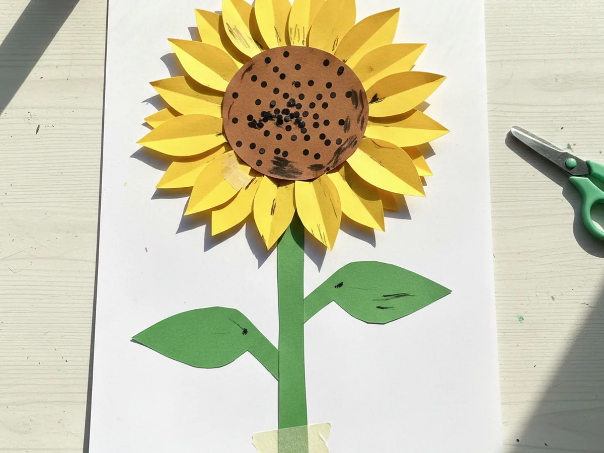Green paper stem and two leaf shapes glued below the yellow and brown sunflower on white cardstock, completing the sunflower paper craft